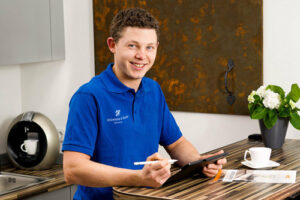 Marius Recker - Junger Mann mit braunen Haaren und blauem Poloshirt sitz mit Tablet am Tisch und lächelt