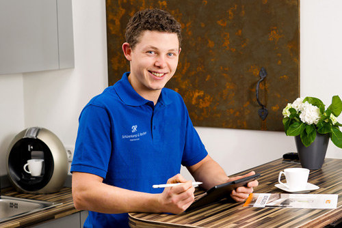 Marius Recker - Junger Mann mit braunen Haaren und blauem Poloshirt sitz mit Tablet am Tisch und lächelt